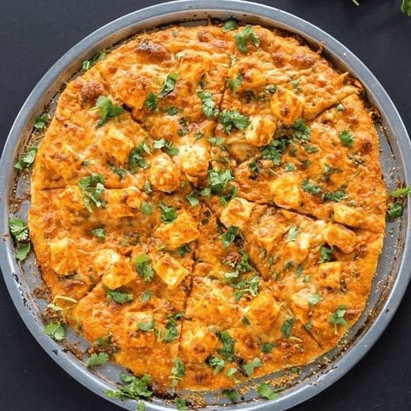 A freshly baked, sliced Pizza Station pizza with paneer and cilantro on a metal pan.
