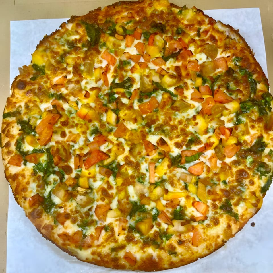 A whole Pizza Station pizza, topped with melted cheese, green pesto, and diced orange and yellow vegetables.