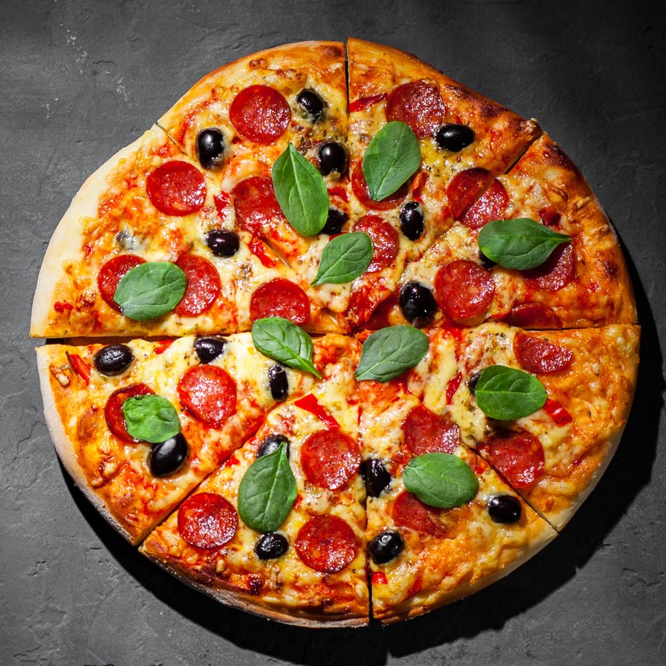 A Pizza Station pepperoni pizza, topped with black olives and fresh basil, sliced on a dark surface.